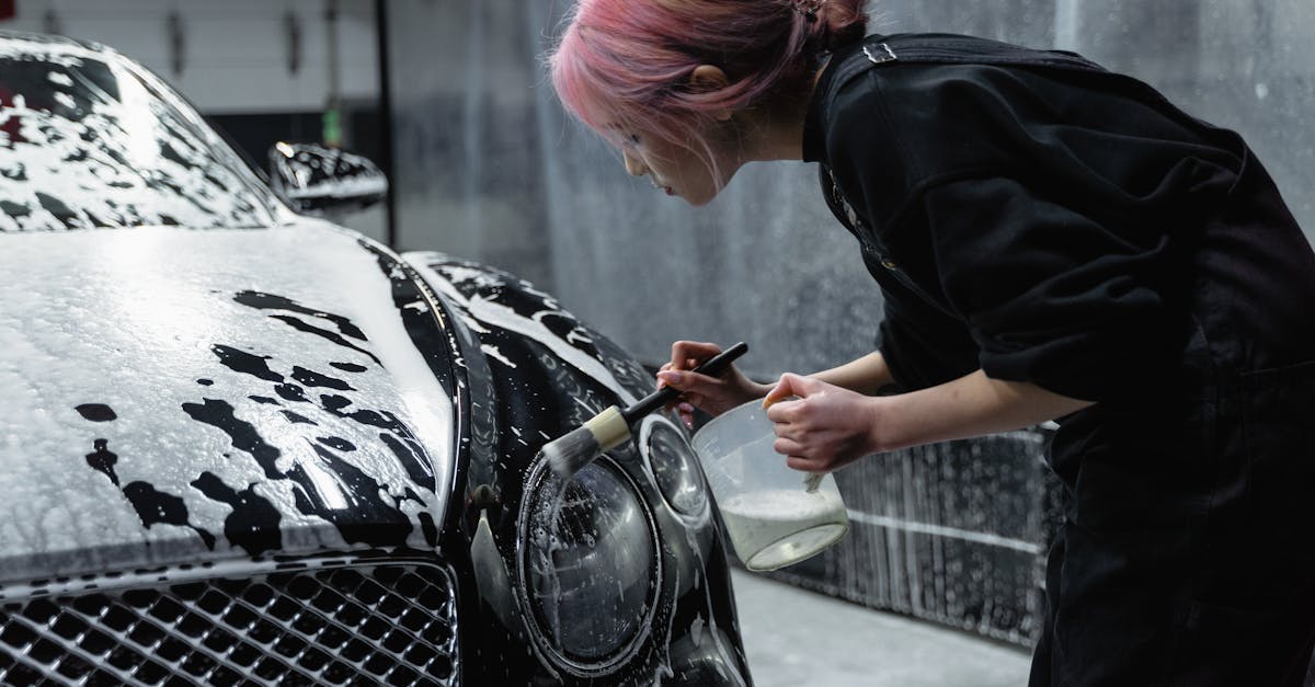 A woman with pink hair meticulously cleans a black car's headlight at an indoor car wash.